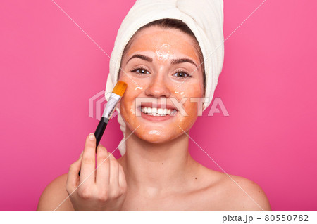 Happy smiling European lady applies cleansing mask with cosmetic brush, has toothy smile, wears white towel, has naked body, poses over pink background, enjoys softness of skin. Skin care concept 80550782