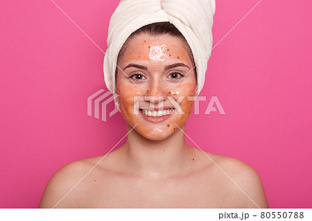 Horizontalshot of beautiful girl wrapped hair with white towel, skin covered by mask. Model with mask on face, posing with bare shoulders, standig indoor against pink studio wall. Beauty concept. Horizontalshot of beautiful girl wrapped hair with white towel, skin covered by mask. Model with mask on face, posing with bare shoulders, standig indoor against pink studio wall. Beauty concept. 80550788