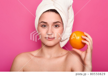 Beauty portrait of young girl with nacked shoulders. Lady holds orange, has calm facial expression. Charming female with towel wrapped around head relaxes in spa salon. Model poses in photo studio. 80550829