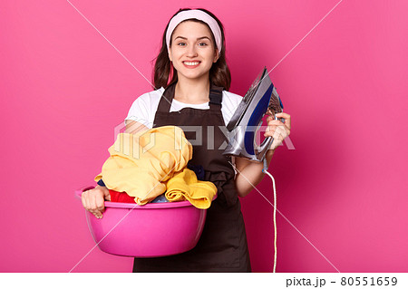 Portrait of cheerful sincere housewife looking directly at camera, wearing brown apron, white headband and t shirt, holding pink basin with laundry and iron, ready to do house chores. Household. Portrait of cheerful sincere housewife looking directly at camera, wearing brown apron, white headband and t shirt, holding pink basin with laundry and iron, ready to do house chores. Household. 80551659
