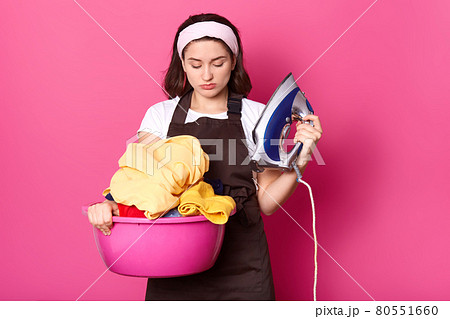 Displeased attractive young female standing isolated over pink background in studio, holding iron and basin with clean clothes in both hands, looking at laundry with disappointment. Householding. Displeased attractive young female standing isolated over pink background in studio, holding iron and basin with clean clothes in both hands, looking at laundry with disappointment. Householding. 80551660