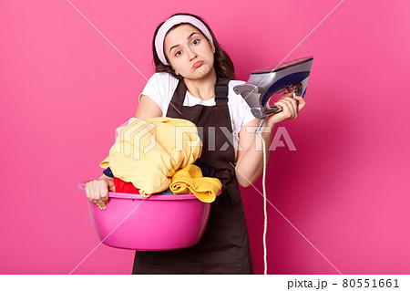 Pretty young woman housewife ready for ironing clean washed things, has broken iron, holds pink basin with fresh linen, squeezes shoulders, wears hair band, casual t shirt, apron, isolated on rose. Pretty young woman housewife ready for ironing clean washed things, has broken iron, holds pink basin with fresh linen, squeezes shoulders, wears hair band, casual t shirt, apron, isolated on rose. 80551661