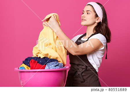 Profile of delighted attractive brunette holding light shirt after washing, being satisfied with result, smiling sincerely, posing isolated over pink background in studio. Housewife lifestyle concept. Profile of delighted attractive brunette holding light shirt after washing, being satisfied with result, smiling sincerely, posing isolated over pink background in studio. Housewife lifestyle concept. 80551689