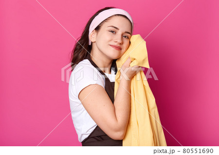 Pleasant charismatic female standing isolated over pink background in studio, wearing brown apron, white t shirt and headband, pressing yellow shirt to her body, enjoying results of washing. Pleasant charismatic female standing isolated over pink background in studio, wearing brown apron, white t shirt and headband, pressing yellow shirt to her body, enjoying results of washing. 80551690