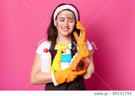 Positive Caucasian woman speaking on mobile phone while clening her house, holding sponge and detergents in hands, dressed in casual clothes, wears rubber gloves and apron, isolated over pink wall. 80551744
