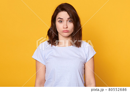Studi oshot of dark haired young woman dresses casual white t shirt, standing and looking at camera, posing with pouty lips isolated over yellow background, being offended and having bad mood. 80552686