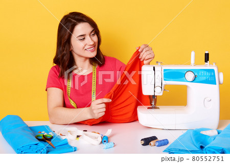 Close up portrait of brunette European female seamstress with tapes mesure on neck, wears casual red t shirt, isolated over yellow concrete wall. Needles, scissors, threads, sewing machine on table. 80552751