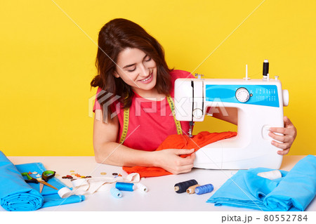 Horizontal picture of hard working professional seamstress sewing item of clothes with help of sewing machine, getting pleasure, smiling sincerely, sitting at table, isolated over yellow background. Horizontal picture of hard working professional seamstress sewing item of clothes with help of sewing machine, getting pleasure, smiling sincerely, sitting at table, isolated over yellow background. 80552784