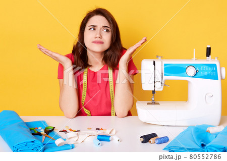 Studio shot of tsylist girl artisan seamstress sitting at white desk with sewing machine, stitch, needles. Attractive brunette sewer looks aside and shrugging, has problems with her equipment. 80552786