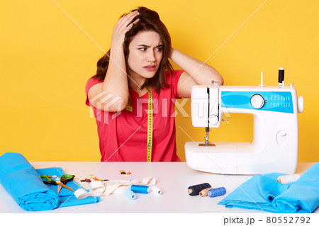 Indoor shot of seamstress sewing in studio, keeps hands on had, has broken sewing machine, wearing red t shirt, has measure tape on neck, surrounded by different sewing equipment, isolated on yellow. 80552792