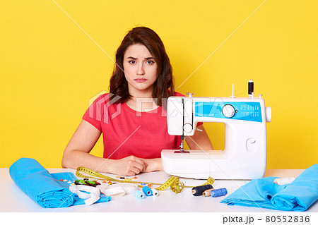 Close up portrait of sad seamstress working at her workshop, adorable dressmaker wearing red casual t shirt, surrounded different equipments, looks upset, has mistake, isolated over yellow background. Close up portrait of sad seamstress working at her workshop, adorable dressmaker wearing red casual t shirt, surrounded different equipments, looks upset, has mistake, isolated over yellow background. 80552836