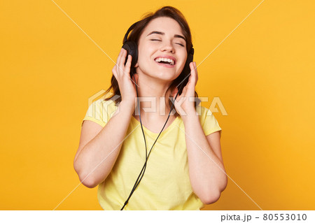 Beautiful woman listening to music on yellow background. Charming lady posing with closed eyess, enjoys of listening favourite music, keeps hands on headphones, sings and relaxes. Lifestyle concept. Beautiful woman listening to music on yellow background. Charming lady posing with closed eyess, enjoys of listening favourite music, keeps hands on headphones, sings and relaxes. Lifestyle concept. 80553010
