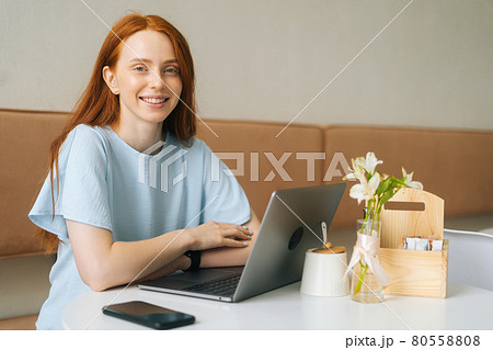 Portrait of cheerful attractive young woman sitting at table with laptop and phone in cafe with warm daylight and looking at camera. 80558808