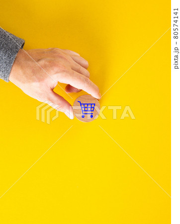 Male hand placing a blue shopping cart icon on yellow background. Male hand placing a blue shopping cart icon on yellow background. 80574141