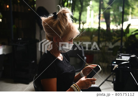 Blonde woman, customer at beauty salon, wearing medical mask, and using mobile phone for work 80575112