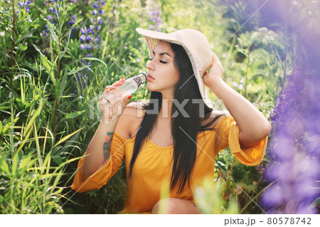 Woman in straw hat and yellow dress drinks water from glass bottle in field of lupins in summer 80578742
