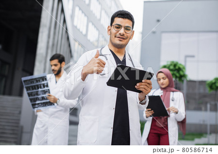 Male doctor with tablet pc, standing with coworkers outdoors near hospital, showing thumb up Male doctor with tablet pc, standing with coworkers outdoors near hospital, showing thumb up 80586714