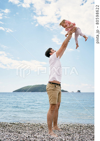 Dad tosses a little girl in his arms while standing on a pebble beach Dad tosses a little girl in his arms while standing on a pebble beach 80598622
