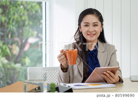 Asian female office manager with brown suit holding tablet computer in her hand. She smile happily at the taste of hot coffee. Morning work atmosphere in a modern office. 80599690