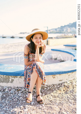 Smiling girl in a hat and dress sits on a bench on a pebble beach Smiling girl in a hat and dress sits on a bench on a pebble beach 80602201