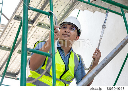 During work at a height, a construction worker wears a safety harness belt. on a background of a drilling platform for a structural rig During work at a height, a construction worker wears a safety harness belt. on a background of a drilling platform for a structural rig 80604043