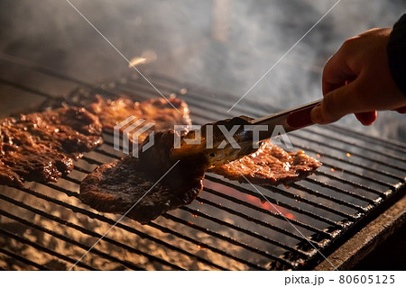 in the evening a barbecue grill on which tasty juicy steaks are grilled over an open fire, a hand holds tongs and turns the meat over. close-up, soft focus. smoke is highlighted 80605125