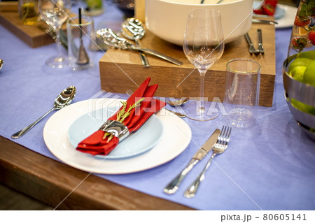 table setting with a blue tablecloth, a white and blue plate, a red napkin in a steel holder with a branch of rosemary, steel appliances, glass goblets. close up, soft focus 80605141