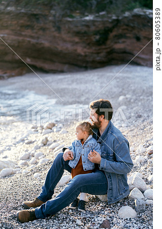 Dad sits on a stone and hugs a little girl on a pebble beach against the backdrop of rocks. Daughter and dad are looking at the sea. Side view 80605389
