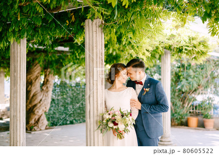 Smiling groom hugs the bride's shoulders against the background of columns in a green garden Smiling groom hugs the bride's shoulders against the background of columns in a green garden 80605522
