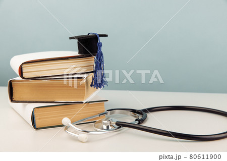 Medical education concept. Stethoscope and graduation cap on stack of books on blue background Medical education concept. Stethoscope and graduation cap on stack of books on blue background 80611900