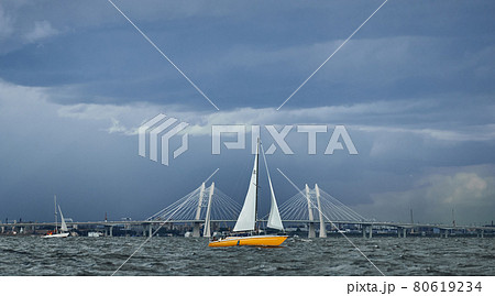 The yellow sailboat on the horizon in sea at sunset, cable bridge on background, the storm sky of different colors, big waves, sail regatta, cloudy weather 80619234