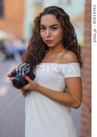 Young attractive female student or tourist using a mirrorless camera while walking in summer city. Woman takes pictures and enjoy weather. Young attractive female student or tourist using a mirrorless camera while walking in summer city. Woman takes pictures and enjoy weather. 80619463