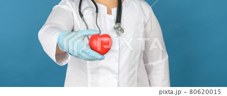 female medic in a white coat stands and holds a red heart on a blue background, the concept of donation and kindness 80620015