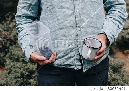 Man in blue linen shirt holding tin cup and plastic container with blueberries in dirty hands. Harvesting foraging wild bilberries fruit. Camping travel concept. Blurred woodland, forest background Man in blue linen shirt holding tin cup and plastic container with blueberries in dirty hands. Harvesting foraging wild bilberries fruit. Camping travel concept. Blurred woodland, forest background 80635901