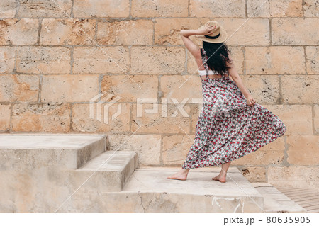 Beautiful barefoot brunette model with long dark hairs, straw hat and floral summer dress posing. Trendy summer fashion lifestyle concept. Concrete stairs and sandstone wall background. Back view. Beautiful barefoot brunette model with long dark hairs, straw hat and floral summer dress posing. Trendy summer fashion lifestyle concept. Concrete stairs and sandstone wall background. Back view. 80635905