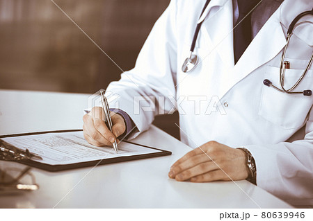 Unknown male doctor sitting and working with clipboard of medication history record in clinic at his working place, close-up. Young physician at work. Perfect medical service, medicine concept 80639946