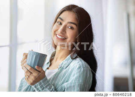 Portrait of pretty Indian lady standing near window with cup of hot drink, smiling at camera, indoors 80640218