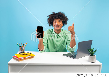 Cheerful black student guy holding smartphone with blank screen and showing thumb up, sitting over blue background Cheerful black student guy holding smartphone with blank screen and showing thumb up, sitting over blue background 80640324