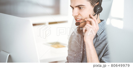 Casual dressed young man using headset and computer while talking with customers online in sunny office. Call center, business concept 80649979