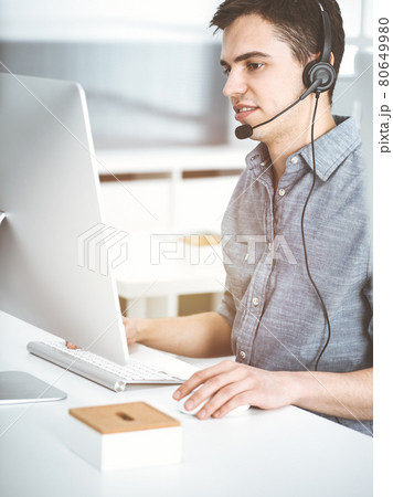 Casual dressed young man using headset and computer while talking with customers online in sunny office. Call center, business concept 80649980