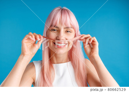 Portrait of happy young woman with natural long pink dyed hair holding a strand of hair as a moustache and smiling at camera, posing isolated over blue studio background 80662529