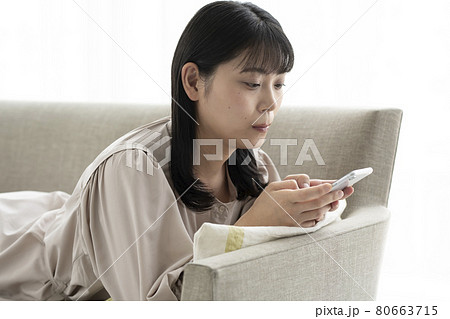 窓際のソファーで寝そべってスマホを操作する女性 80663715