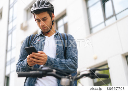 Low-angle view of handsome male courier with backpack standing with bicycle in city street and using navigation app on phone, close-up. 80667703
