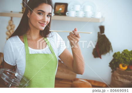 An attractive young dark-haired woman tasting ready hot meal with a wooden spoon while standing and smiling near the kitchen stove. Cooking and householding concepts 80669091