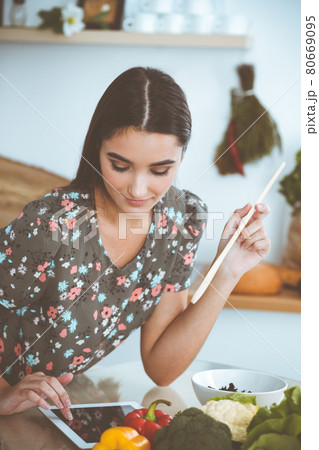 An attractive young dark-haired woman tastes a new recipe for a delicious salad mix while sitting at the table in the kitchen. Tablet pc is the best cookbook 80669095