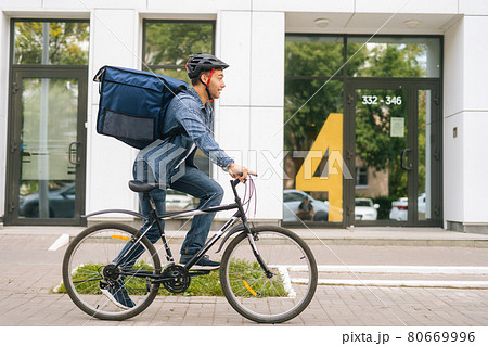 Side view of happy handsome young delivery man in protective helmet starting bike ride in city street on background of office building. Side view of happy handsome young delivery man in protective helmet starting bike ride in city street on background of office building. 80669996