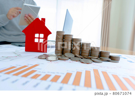 Businessman using a tablet to analyze the data with a laptop computer near the table with piles of coins and house designs, graphs, and glasses. Investment concept. Businessman using a tablet to analyze the data with a laptop computer near the table with piles of coins and house designs, graphs, and glasses. Investment concept. 80671074