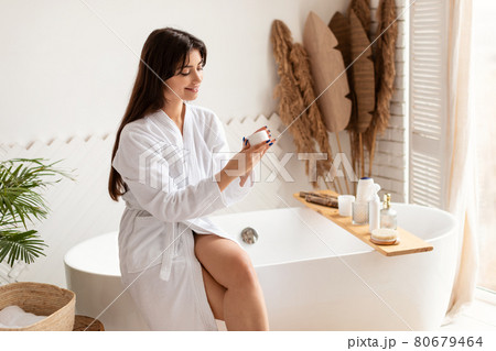 Cheerful Brunette Woman Holding Cream Moisturizer Sitting In Modern Bathroom Cheerful Brunette Woman Holding Cream Moisturizer Sitting In Modern Bathroom 80679464