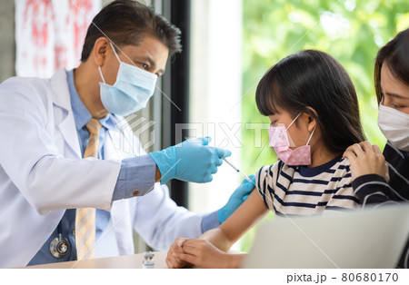 Asian senior doctor wearing gloves and isolation mask is making a COVID-19 vaccination in the shoulder of child patient with her mother at hospital. Asian senior doctor wearing gloves and isolation mask is making a COVID-19 vaccination in the shoulder of child patient with her mother at hospital. 80680170