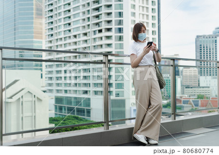 solo asian woman wear protective mask during use phone and relax at rooftop with city background 80680774